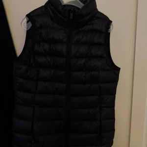 Svart dunväst med dragkedja - En klassisk svart dunväst med hög krage och dragkedja framtill. Västen är quiltad och har en stilren, sportig look som passar perfekt till lager-på-lager-stil.