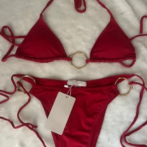 Röd bikini med guldringar från Museara - Trendig röd bikini från Museara med guldfärgade ringdetaljer både på överdelen och nederdelen. Klassisk trekantsmodell med knyt i sidorna och i nacken. Perfekt för sommaren!