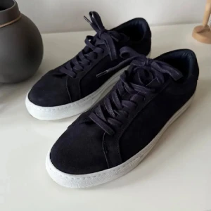 Marinblåa sneakers i mocka från Les Deux - Snygga marinblåa sneakers från Les Deux i mocka med vita sulor. Skorna har snörning och en diskret detalj med röd och vit rand på hälen. Perfekta för en stilren look. De är knappt använda någonting. Storlek 42. Nypris 1499kr