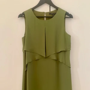Olivgrön ärmlös klänning från BCBG Max Azria - Säljer en olivgrön ärmlös klänning från BCBG Max Azria. Klänningen har en elegant lager-på-lager design framtill och en rak, enkel siluett. Perfekt för dig som gillar stilrena och moderna plagg.