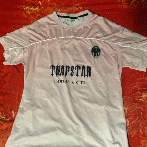 Rosa fotbollströja från Trapstar - Säljer en rosa fotbollströja från Trapstar med svart tryck på bröstet och ryggen. På framsidan står det 'Trapstar It's a secret' och på ryggen finns stort nummer 22. Tröjan har korta ärmar och rund hals.