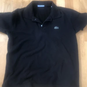 Svart pikétröja från Lacoste - Klassisk svart pikétröja från Lacoste med broderad logga på bröstet och ärmen. Tröjan har krage, korta ärmar och knappar vid halsen. Perfekt för en stilren och sportig look.