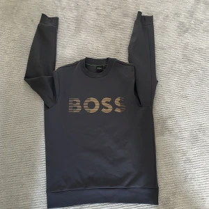 Svart sweatshirt från BOSS - Säljer en svart sweatshirt från BOSS med guldigt logotryck framtill. Tröjan har rund halsringning och långa ärmar. Perfekt för en stilren och avslappnad look.( Inga prutningar)
