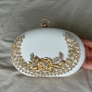 Vit clutch/kuvertväska med gulddetaljer  - Helt ny! Etikett finns kvar. Superfin oval kuvertväska i vitt med guldfärgad kedja och dekorativa diamanter samt blomma framtill. Väskan har ett elegant metallspänne och kedja som axelrem. Perfekt för speciella tillfällen som bröllop, bal, fest osv. 