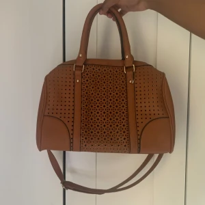 Brun handväska med utskurna detaljer - Säljer en snygg brun handväska med dekorativa utskurna mönster på framsidan. Väskan har både handtag och en avtagbar axelrem, guldfärgade metalldetaljer och mörkblått interiör. Har aldrig använts. 