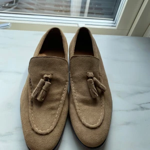 Beige mocka loafers med tofsar - Säljer ett par stilrena beige loafers i mocka med dekorativa tofsar framtill. Skorna har rund tå och platt sula, perfekta för en dressad look. Klassisk design som passar till många tillfällen.