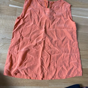 Orange/persikofärgad  ärmlös linnetopp i siden - Säljer en orange ärmlös linnetopp i 100% siden. Toppen har rund halsringning och en liten knapp i nacken. Perfekt för varma dagar och enkel att matcha med olika nederdelar. Obs! Första och andra bilden visar nyansen på toppen. De andra bilderna är tagna i ett ljus som får toppen att se mer orange ut. 