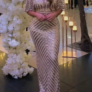 Glittrig långklänning i guld - Säljer en glittrig långklänning i guld med paljetter och kort ärm. Klänningen har figurnära passform och ett elegant mönster av diagonala ränder. Perfekt för festliga tillfällen och bal.