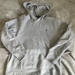 Grå hoodie från Polo Ralph Lauren - Säljer en klassisk grå hoodie från Polo Ralph Lauren med broderad logga på bröstet. Tröjan har huva, magficka och långa ärmar. Perfekt för en avslappnad och stilren look.