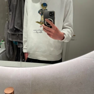 Vit hoodie från Polo Ralph Lauren med björntryck - Vit hoodie från Polo Ralph Lauren med ikoniskt björntryck på bröstet. Tröjan har huva med dragsko och ribbade muddar vid ärmslut och nederkant. Tillverkad i en mjuk bomullsblandning för extra komfort. Värde på 2000 kr. Första bilden är hoodien nyligen strykt. Kontakta gärna vid fler funderingar.