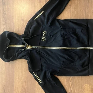 Svart hoodiejacka från BOSS - Svart hoodiejacka från BOSS med gulddetaljer och dragkedja framtill. Jackan har huva, två fickor och dekorativa ränder på ärmarna. Perfekt för en sportig och stilren look.