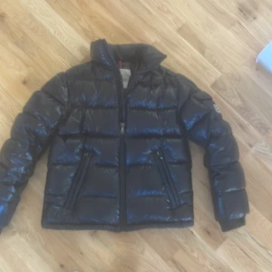 Svart dunjacka från Moncler - Säljer en svart, glansig dunjacka från Moncler med klassisk pufferdesign. Jackan har dragkedja framtill, två snedställda fickor med dragkedja och lång ärm. Perfekt för kyliga dagar och riktigt snygg streetstil.