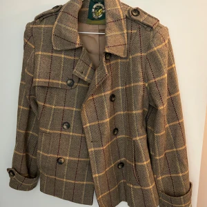 Rutig kavajjacka från Oxford Blue - Rutig kavajjacka i beige och bruna toner med gula och röda ränder. Jackan har knappar framtill, axelklaffar och fickor på bröstet. Klassisk krage och dekorativa knappar vid ärmslut. 