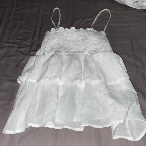 Vit volanglinnetopp från Gina Tricot - Säljer en söt vit linnetopp från Gina Tricot med smala axelband och volangdetaljer. Toppen har smock upptill och är luftig med flera lager tyg, perfekt för varma dagar. Jättefin topp som tyvärr aldrig har använts. Den är i nyskick med inga slitningar.