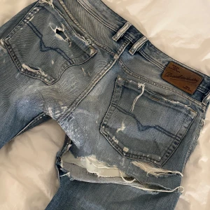 Blå slitna jeansbyxor från Diesel - Säljer ett par blå jeansbyxor från Diesel med tydliga slitningar och hål, särskilt på bakfickor och lår. Klassisk femficksmodell med knapp och dragkedja. Perfekt för dig som gillar en rå och avslappnad stil.