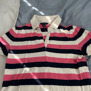 Randig pikétröja från Tommy Hilfiger - Säljer en klassisk pikétröja från Tommy Hilfiger med breda ränder i rosa, marinblått och vitt. Tröjan har krage, knappar framtill och korta ärmar. Perfekt för en sportig och stilren look! Pris kan diskuteras