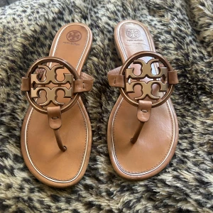 Tory Burch - Säljer ett par snygga bruna sandaler från Tory Burch med stort logodetalj i metall framtill. Skorna har platt sula och öppen tå, perfekta för varma dagar. Klassisk och stilren design med läderremmar. Använda ett fåtal gånger. Nypris 3400kr