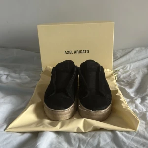 Svarta sneakers från Axel Arigato - Säljer ett par svarta sneakers från Axel Arigato med tjock beige sula. Skorna har en stilren design utan snörning och är tillverkade i mocka. Perfekta för dig som gillar minimalistisk stil och vill ha något som sticker ut. Finns mycket slitningar varav det låga priset.