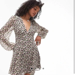 Blommig kortklänning från Topshop - Superfin kortklänning från Topshop med svart botten och blommigt mönster i rosa och grönt. Klänningen har v-ringning, långa transparenta ärmar och lätt utsvängd kjol. Perfekt för dig som gillar romantisk stil! Aldrig använt och prislapp är kvar:)