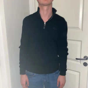 Svart tröja med halv dragkedja från Superdry - Säljer en svart långärmad tröja från Superdry med halv dragkedja vid halsen. Tröjan har en klassisk passform och är tillverkad i mjukt material, perfekt för vardagsbruk. Diskret logga på bröstet. Storleken är s men den passar även storleken m. Han på bilden är 180 och väger 62 