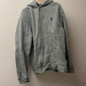 Polo Ralph Lauren hoodie - Säljer en klassisk grå hoodie från Polo Ralph Lauren med dragkedja och fickor framtill. Tröjan har broderad logga på bröstet och är tillverkad i mjukt material. Perfekt för en avslappnad stil. Tar inte emot byten. Det är först till kvarn. Barn storlek XL.