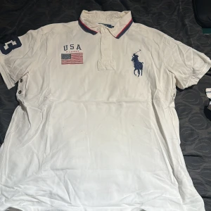 Vit piké t-shirt från Ralph Lauren - Vit piké t-shirt från Ralph Lauren med blå logga på bröstet, USA-flagga och texten 'USA' samt nummer 3 på ärmen. Kragen har röd och marinblå kant. Klassisk och sportig stil.