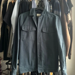 Blå Jacka från These Glory Days - Säljer en stilren jacka från These Glory Days. Skjortan har två bröstfickor med lock och knappar samt klassisk krage. Tillverkad i ett kraftigare bomullstyg, perfekt som lager över t-shirt.