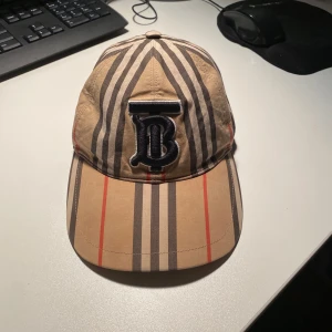 Beige rutig keps från Burberry - Säljer en riktigt fin beige keps från Burberry med deras ikoniska rutiga mönster och ett stort svart broderat B framtill. Kepsen har justerbar rem baktill och är tillverkad i bomull. Kepsen sen skrynklig ut på bild, men försvinner så fort man tar på sig den. Priset är diskutabelt!