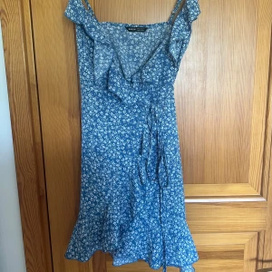 Blå blommig omlottklänning - Söt blå omlottklänning från shein med vitt blommönster. Klänningen har volangdetaljer, knyt i midjan och är ärmlös med smala axelband. Perfekt för varma dagar och somriga tillfällen.