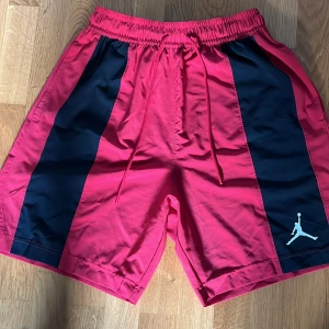 Röd svarta Jordan shorts - Säljer ett par snygga röda shorts från Jordan Nike med svarta paneler på sidorna och vit Jordan logo. Shortsen har elastisk midja med snörning och är perfekta för sport eller chill. Materialet känns lätt och luftigt.
