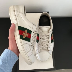 Gucci ace skor - Säljer ett par klassiska vita sneakers från Gucci med grönt och rött band på sidan och en broderad guldfärgad bi. Skorna har snörning och rund tå. Perfekta för dig som gillar lyxiga detaljer och stilren design. Storlek 42 men passar 43. Box tillkommer. Skorna är finare skick i verkligheten.