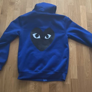 Blå hoodie från Comme des Garçons Play - Blå hoodie med stor svart hjärtformad detalj och ögon på ryggen från Comme des Garçons Play. Klassisk huva och ribbade muddar. Perfekt för dig som gillar streetwear och vill sticka ut.