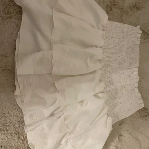 Vit volangkjol  - Supersöt vit kjol med bred smock i midjan och flera volanglager. Kjolen är kort och har en luftig känsla, perfekt för varma dagar. Enkel att matcha med olika toppar för en fräsch look.