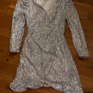 Beigeglittrig omlottklänning - Säljer en beigefärgad glittrig omlottklänning med långa ärmar och v-ringning. Klänningen har en snygg slits framtill och är täckt av paljetter som ger ett riktigt festligt intryck. Perfekt för dig som vill sticka ut på nästa event!