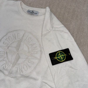 Vit stickad tröja från Stone Island - Vit stickad tröja från Stone Island med stort broderat logomärke på bröstet. Klassisk rund halsringning och ribbade muddar vid ärmslut och nederkant. 