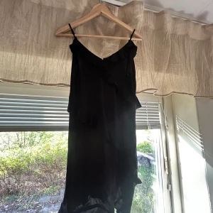 Svart omlottklänning med volang - Säljer en svart omlottklänning med smala axelband och volangdetaljer runt hela klänningen. Klänningen har en elegant v-ringning och är tillverkad i ett lätt, mjukt material som faller fint.