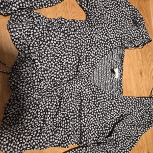 Svart blommig topp från H&M - Säljer denna svarta tunika från H&M med vita småblommor. Toppen har lång ärm med resår vid ärmslut och v-ringning. Materialet är mjukt och stretchigt, perfekt för vår och sommar.