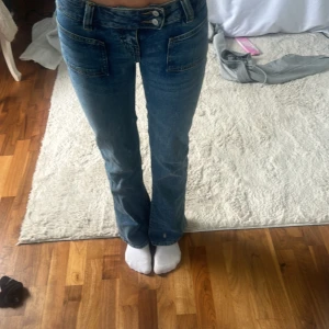 Blå bootcut jeans med dubbla knappar - Säljer ett par blå bootcut jeans med dubbla knappar i midjan och stora fickor fram. Jeansen har en klassisk tvätt och är lågmidjade. Perfekta för dig som gillar retrostil och vill ha ett par snygga jeans till vardags.
