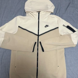 Nike tech  - Snygg beige och vit hoodie från Nike med svart dragkedja och logga på bröstet. Passar väldigt bra för mig som är 180 och är andvänd ett par gånger efter köp, säljer för ett rimligt pris gemfört med nypris🤝
