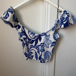 Blåvit blommig croppad topp från H&M - Säljer en blåvit croppad topp från H&M med blommigt mönster. Toppen har offshoulder-modell och använd 1 gång