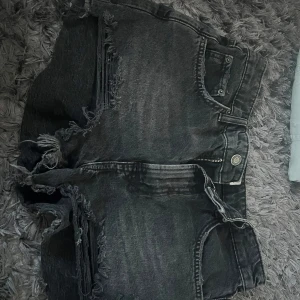 Shorts - Säljer ett par svarta jeansshorts från Zara med råa franskantade benslut och slitna detaljer. Shortsen har klassisk femficksdesign, hög midja och dragkedja med knapp. Perfekta för en cool och avslappnad stil.