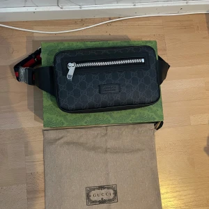Svart axelväska från Gucci med monogram - Säljer en svart axelväska från Gucci med klassiskt GG-monogram och silverfärgad dragkedja. Väskan har justerbart band i rött och blått och levereras med originalpåse och låda. Perfekt för att bära det viktigaste med stil.