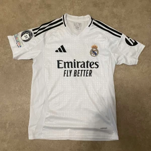 Real Madrid 24/25 VINI JR. #7 - Säljer en vit Real Madrid hemmatröja från Adidas med Vini Jr och nummer 7 på ryggen. Tröjan har korta ärmar, svarta detaljer och klubbmärke broderat på bröstet. Sponsorlogga på framsidan och Champions League-märke på ärmen. Den är nästan äkta