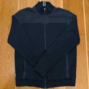 Mörkblå zip-jacka från Hugo Boss - Snygg mörkblå zip-jacka från Hugo Boss med hög krage och diskret broderad logga på bröstet. Jackan har hel dragkedja, ribbade muddar och två sidofickor. Perfekt för en stilren och avslappnad look.