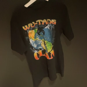 Svart Wu-Tang Clan t-shirt - Cool svart t-shirt med färgglatt Wu-Tang Clan-tryck på framsidan. Perfekt för fans av gruppen som vill ha en avslappnad stil. T-shirten har en loose fit och är kortärmad.