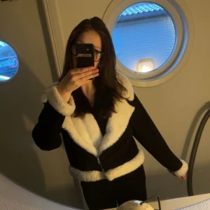 Svart jacka med vit pälsdetalj - Snygg svart jacka med vit pälsdetalj på krage, ärmslut och midja. Jackan har en dragkedja framtill och ger en stilren look. Perfekt för kyligare dagar. Väldigt tunn