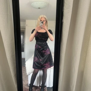 Svart klänning med rosa broderi från Lisa Charmem - Snygg svart klänning med rosa broderade detaljer. Klänningen har korta puffärmar och en smickrande passform. Perfekt för en kväll ute eller en speciell tillställning. 💃(eget moccaskärp)