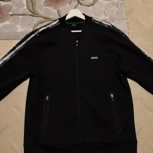 Svart kofta från Boss - Säljer en stilren svart hoodie från Boss med dragkedja och vita detaljer längs ärmarna. Jackan har en enkel design med två fickor framtill och Boss-loggan på bröstet. Perfekt för en casual look.