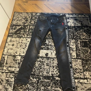 Svarta jeans från Dsquared2 - Snygga svarta jeans från Dsquared2 med coola slitningar och en vit logotyp längs sidorna. De har en klassisk femficksdesign och en röd detalj på framsidan. Perfekta för en trendig look!