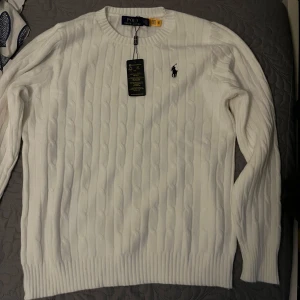 Vit kabelstickad tröja från Ralph Lauren - Säljer en stilren vit kabelstickad tröja från Ralph Lauren. Tröjan har en klassisk rund halsringning och det ikoniska broderade logotypen på bröstet. Perfekt för en elegant och avslappnad look.